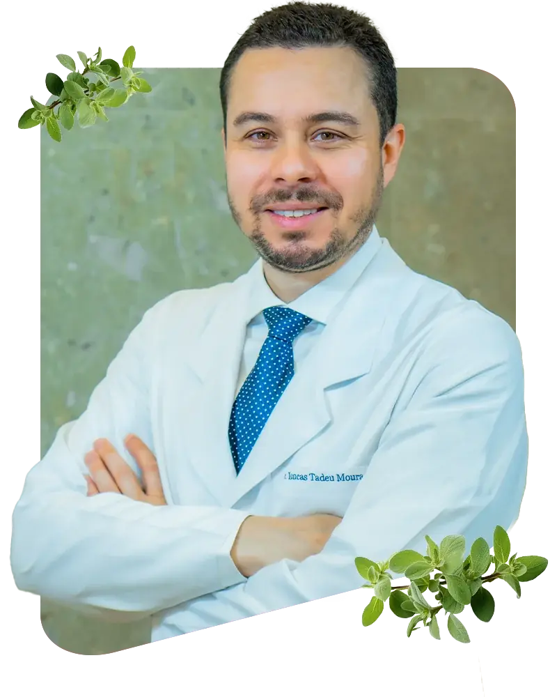 Dr. Lucas Tadeu Moura de jaleco branco e gravata, em pé no consultório, ao lado de um quadro